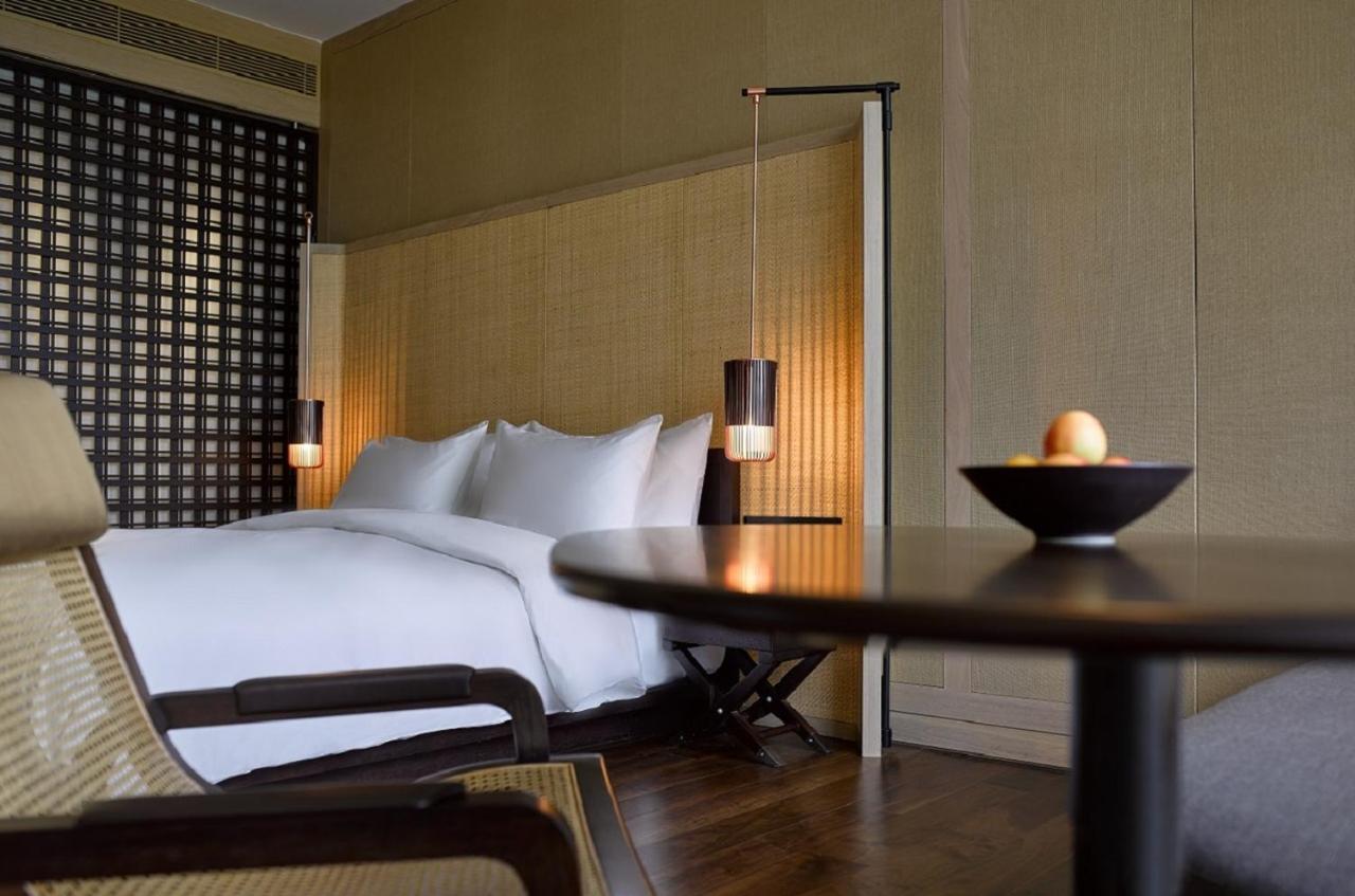 The Ruma Hotel And Residences Kuala Lumpur Kültér fotó The photo shows a well-designed bedroom with a modern and minimalist aesthetic. The room features a large bed with white linens, positioned against a textured wall. On either side of the bed, there are pendant lights that provide a warm glow. The dec