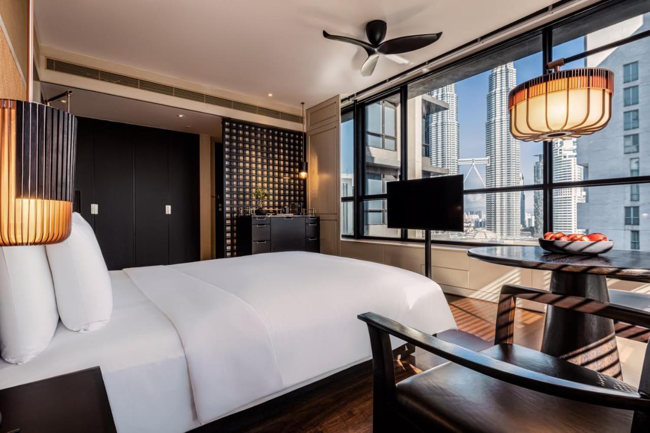 The Ruma Hotel And Residences Kuala Lumpur Kültér fotó The photo shows a modern hotel room featuring a large bed with white linens. There's a ceiling fan above and large windows letting in natural light, providing a view of a cityscape outside. To one side, there's a small seating area with a round table